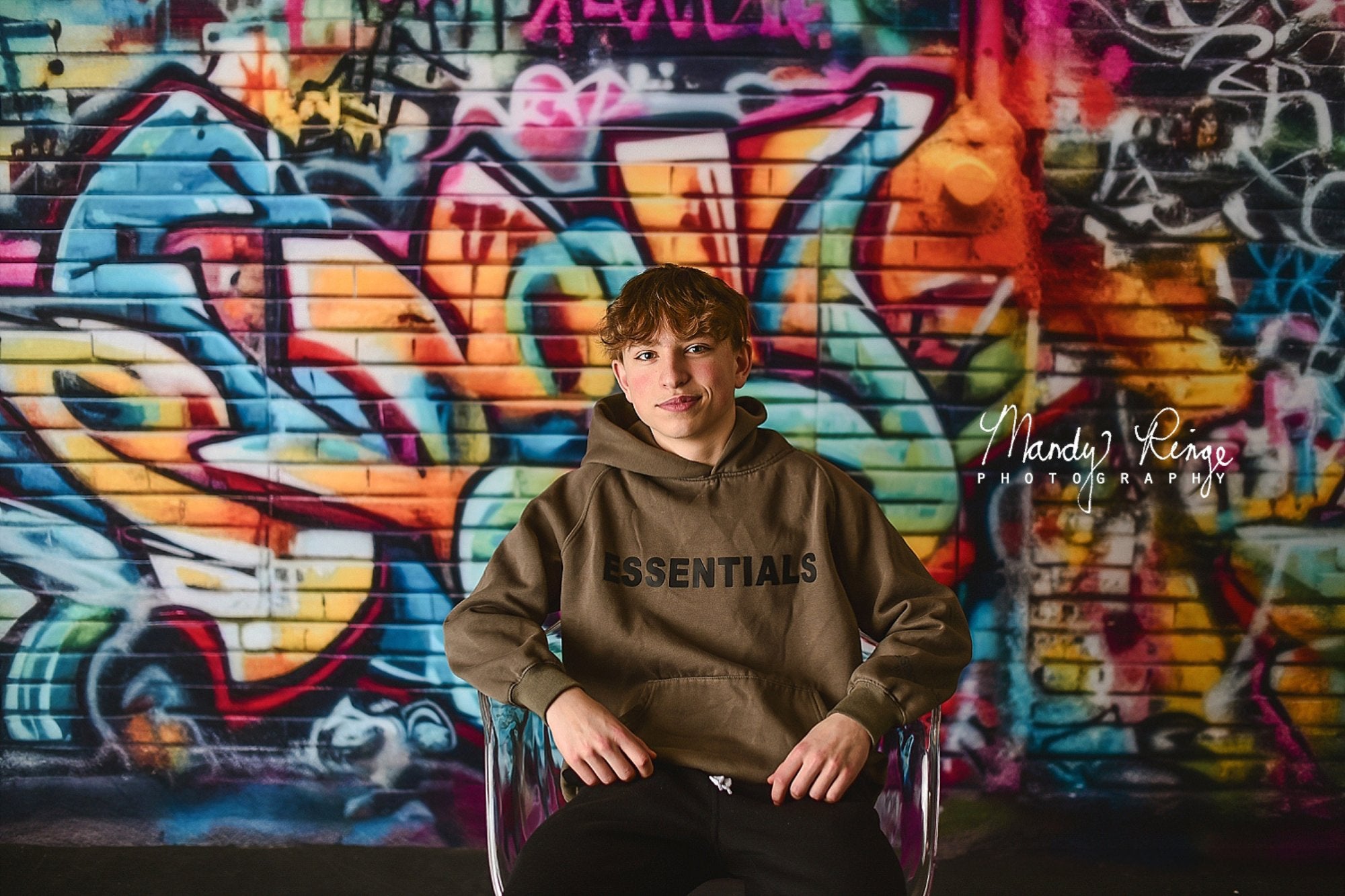 Un garçon est assis sur une chaise transparente devant un fond de mur de briques graffiti coloré