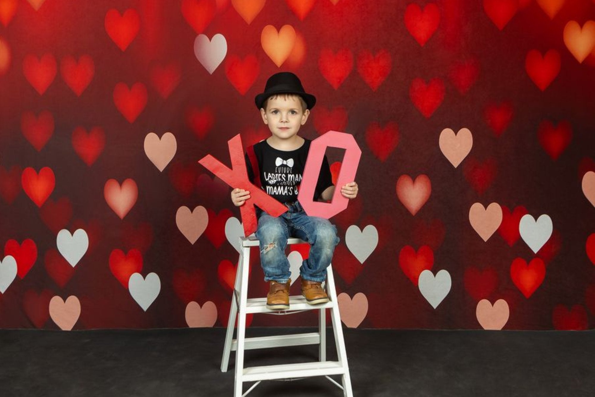 Un garçon est assis devant un tissu de fond rouge à motif cœur, tenant dans ses mains des accessoires de lettres X et O